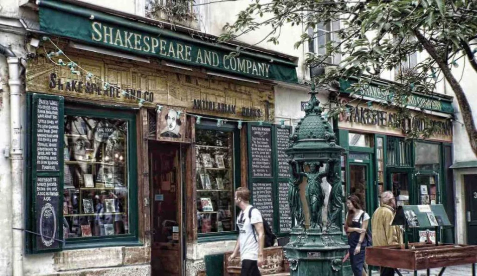 Exterior of Shakespeare and Company bookstore with people passing by and a vintage water fountain.